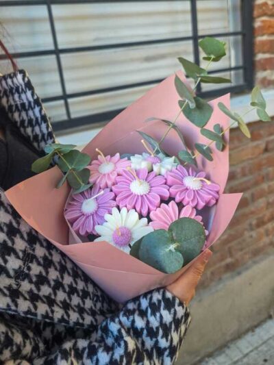 Detalle velas aromáticas de soja en forma de margaritas blancas con centro amarillo y ramas de eucalipto en florero blanco.